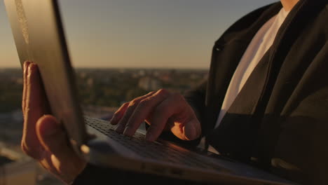 Close-up:-a-programmer's-hand-typing-on-a-laptop-keyboard-at-sunset-overlooking-the-roof.-A-businessman-works-remotely.-Freelancer-performs-work-on-vacation